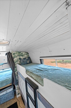 Interior d'una caravana reduït, amb llit superposat, finestra petita i vista cap a l'exterior.