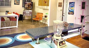 A kitchen with a blender, a dining table with chairs, a small table with a lamp, and a wall with a poster. - Vintage Retro Apartment | Modern Comfort | Cañon City Center (Canon City)