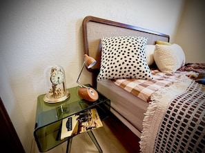 A neatly made bed with a checkered blanket, a polka dot pillow, and a plain pillow. A small glass table beside the bed holds a clock and a lamp. - Vintage Retro Apartment | Modern Comfort | Cañon City Center (Canon City)