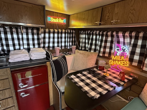 A retro-themed room with a checkered pattern, a neon sign for a snack bar, a milkshakes sign, and a Coca-Cola dispenser. - Vintage Trailer Stay | 1 Unit | Near Ruby's Diner in Cañon City (Canon City)