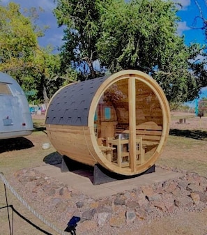 Una construcció de fusta cilíndrica amb una taula i cadires a l'interior, envoltada d'una zona de grava i arbres.