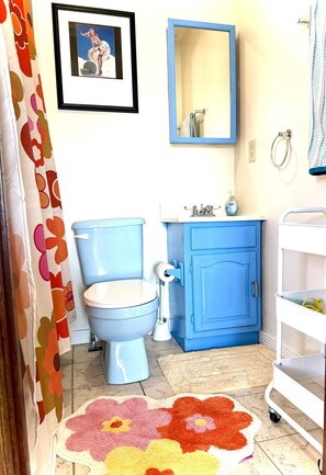 Un baño con inodoro azul, lavamanos, mueble, espejo y cortina de ducha.