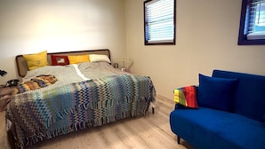 Ein Schlafzimmer mit einem Bett, einem blauen Sofa, einem Fenster mit Jalousien und einem Nachttisch mit Lampe.