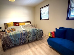 Un dormitorio con cama, sofá azul, ventana con persianas y mesita de noche con lámpara.