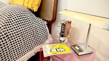 A neatly arranged bedroom with a bed, a bedside table with books, a lamp, and a small table with a magazine.