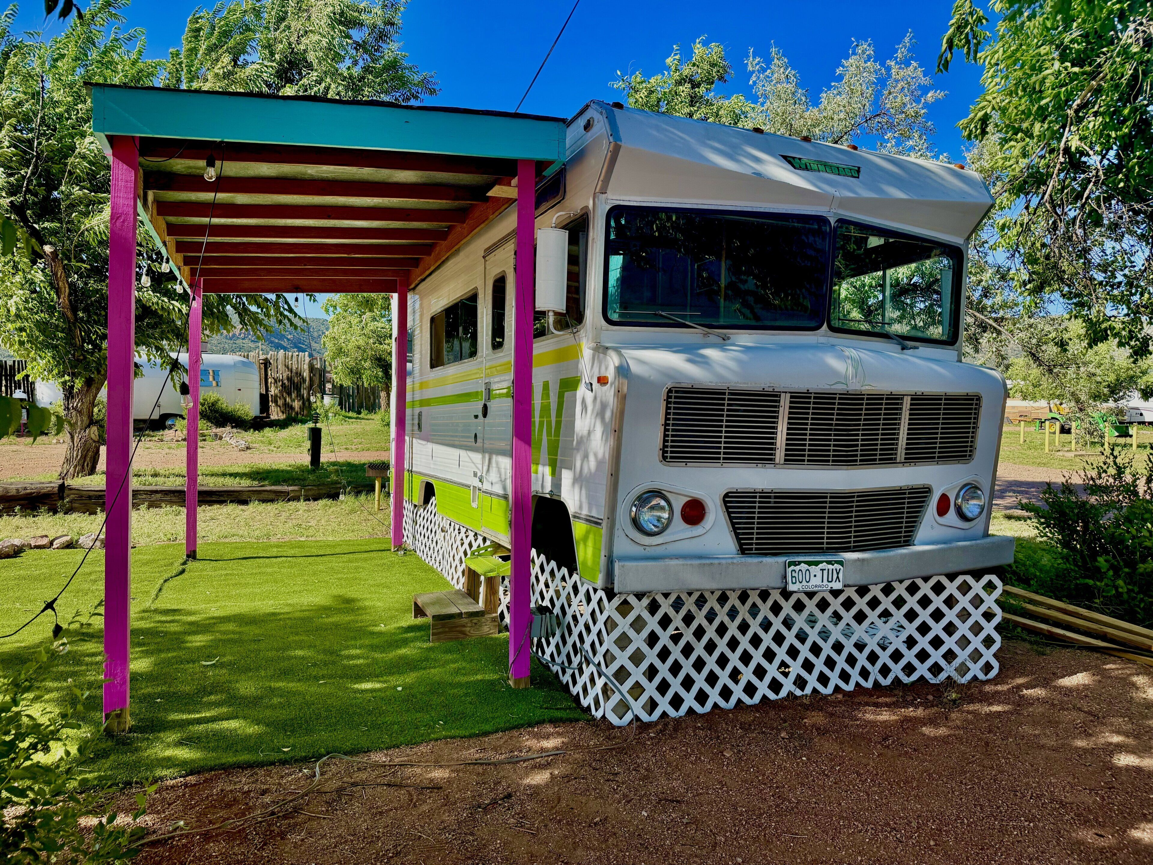 A vintage camper van parked under a covered area with a white lattice base.