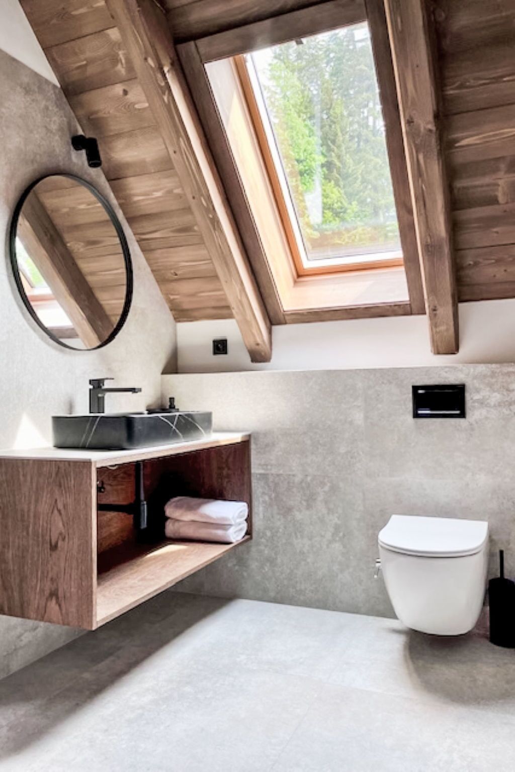Une salle de bain moderne avec un puits de lumière, des poutres en bois, un miroir rond, un lavabo avec un receveur noir et des toilettes blanches.
