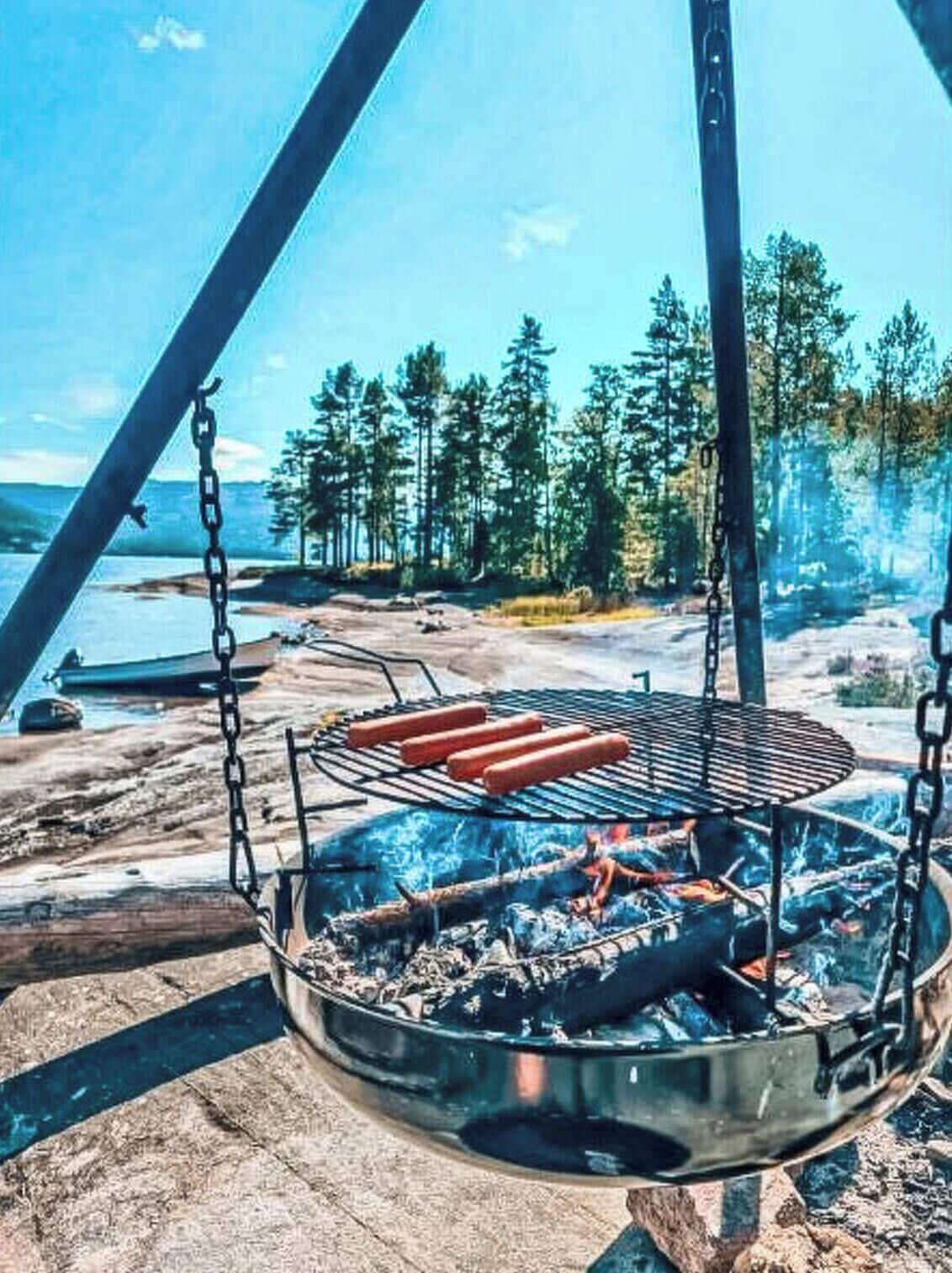 A grill with hot dogs, a fire pit, and a view of a forest and lake.