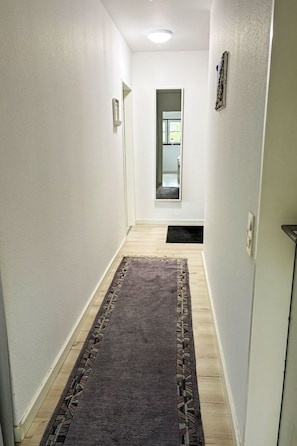 Ein schmaler Flur mit dunklem Teppich, weißen Wänden und einer Tür zu einem Zimmer mit einem Fenster.