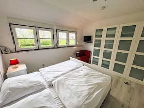 Ein Schlafzimmer mit zwei Betten, einem roten Sessel, einem Fernseher und einem Fenster mit Blick auf Grünflächen.