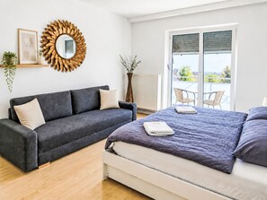 Ein Schlafzimmer mit grauem Sofa, einem Bett mit violettem Überwurf, einem großen Spiegel mit goldener Rahmung und Blick ins Freie durch eine Schiebetür.