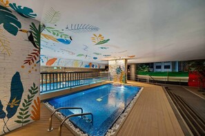 Una piscina interior con un muelle de madera, barandilla de cristal y un mural con motivos de hojas en las paredes.