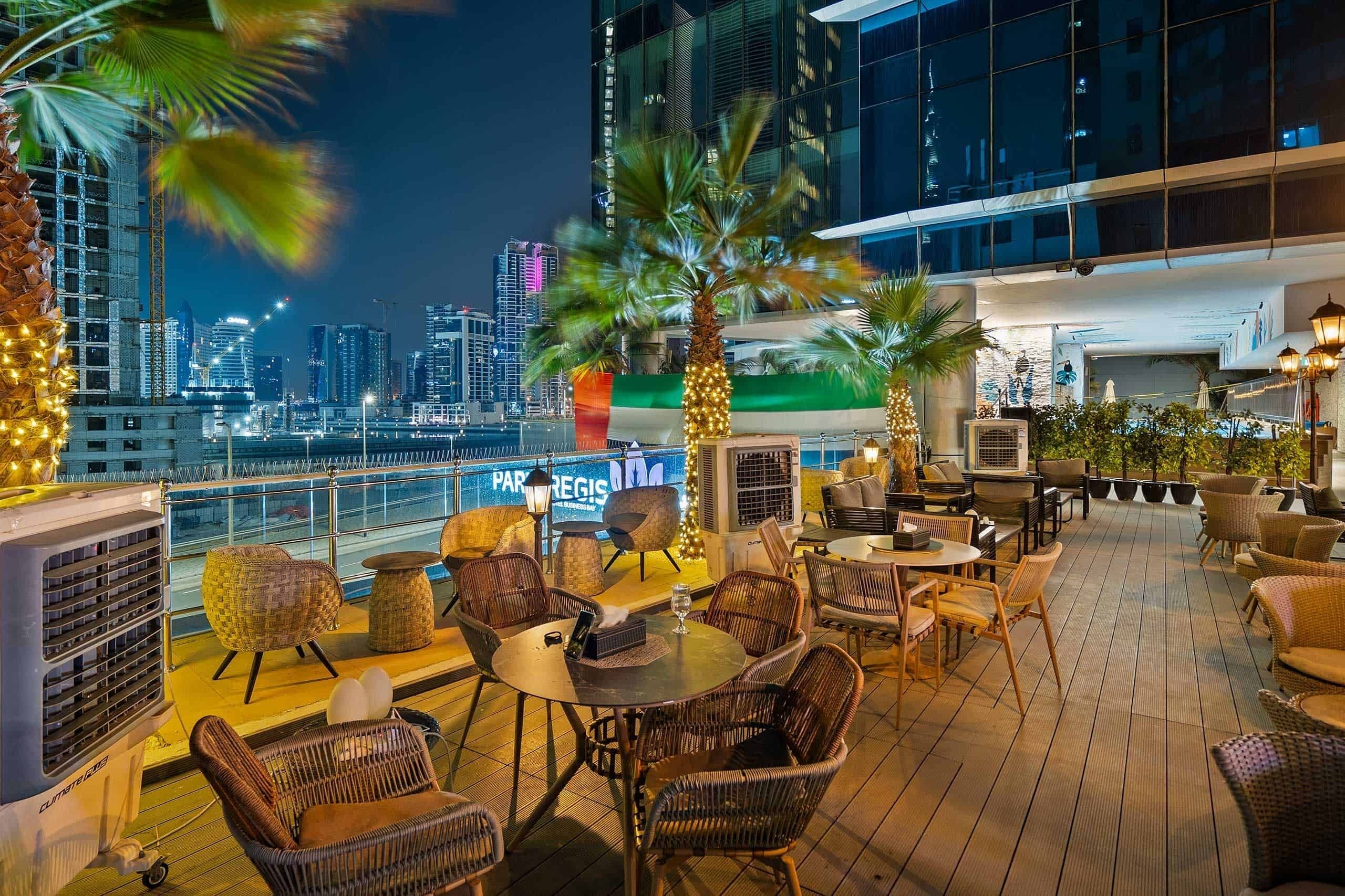 A rooftop bar with wicker furniture, a view of the city skyline, and a sign for 'PAR REGIS.'