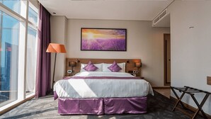 A hotel room with a large bed, a window with curtains, a wall-mounted painting, and a lamp.
