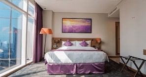 Une chambre d’hôtel avec un grand lit, une fenêtre avec des rideaux, un tableau accroché au mur et une lampe.