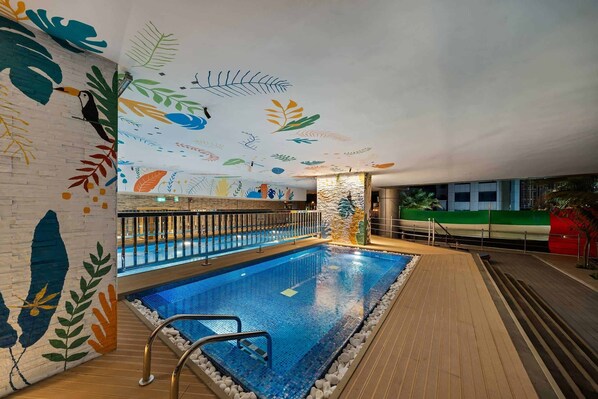 Une piscine intérieure dotée d’une terrasse en bois, d’une balustrade en verre et d’une fresque murale ornée de motifs de feuilles.