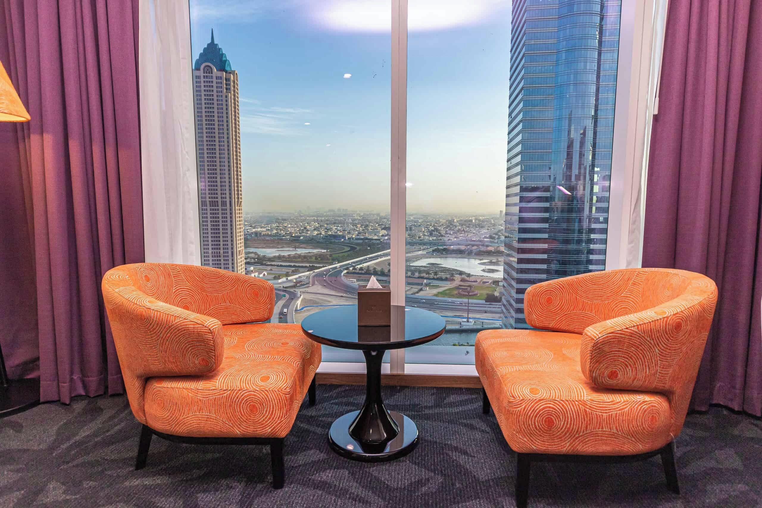 Een hotelkamer met twee oranje fauteuils, een kleine ronde tafel en een groot raam met uitzicht op de stad.