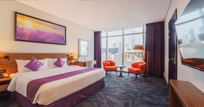 Une chambre d’hôtel avec un grand lit, deux fauteuils, une petite table, un bureau, une lampe, un tableau et une vue sur la ville.