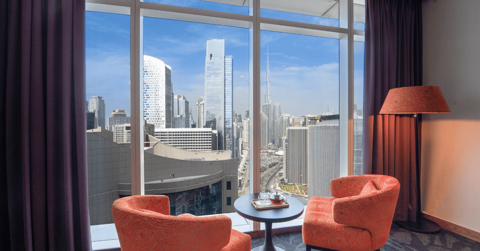 A hotel room with a large window, two orange chairs, a small table, and a lamp.