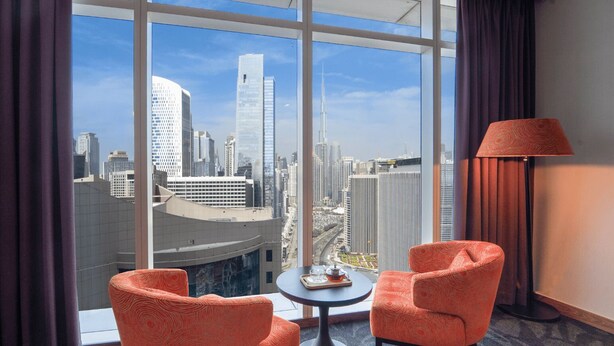 Una habitación de hotel con un ventanal grande, dos sillones anaranjados, una mesita y una lámpara.
