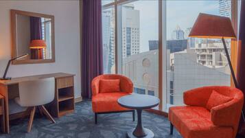 Une chambre d’hôtel équipée d’un bureau, de deux chaises, d’une table ronde, d’un miroir et d’une grande fenêtre donnant sur la ville.