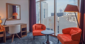 Une chambre d’hôtel équipée d’un bureau, de deux chaises, d’une table ronde, d’un miroir et d’une grande fenêtre donnant sur la ville.
