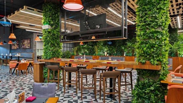 A modern restaurant with a geometric patterned floor, wooden tables and chairs, hanging pendant lights, and green vertical gardens.