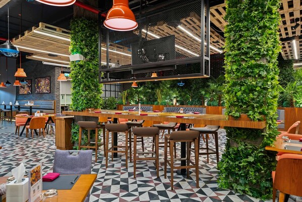 A modern restaurant with a geometric patterned floor, wooden tables and chairs, hanging pendant lights, and green vertical gardens.
