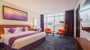 A hotel room with a large bed, two armchairs, a small table, a desk, a lamp, a painting, and a view of the city.