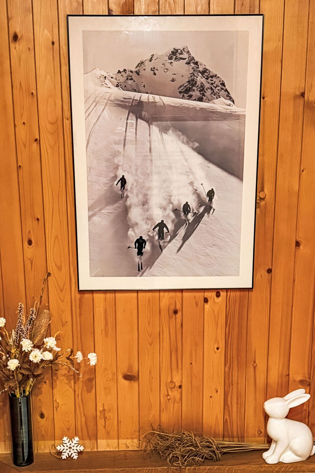 A framed photograph of skiers on a snowy slope, a vase with dried flowers, and a white rabbit figurine on a wooden shelf.