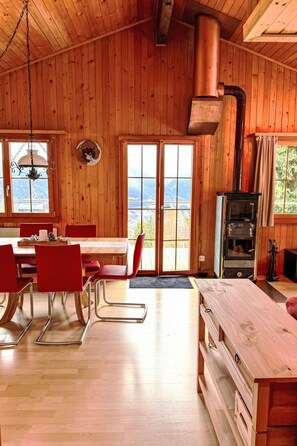 A wooden cabin interior with a dining area, a fireplace, and a view through the large windows. - La Marmotte by Interhome (Bellwald)