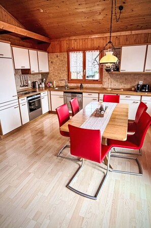 A kitchen with wooden cabinets, a dining table with red chairs, and a hanging light fixture. - La Marmotte by Interhome (Bellwald)