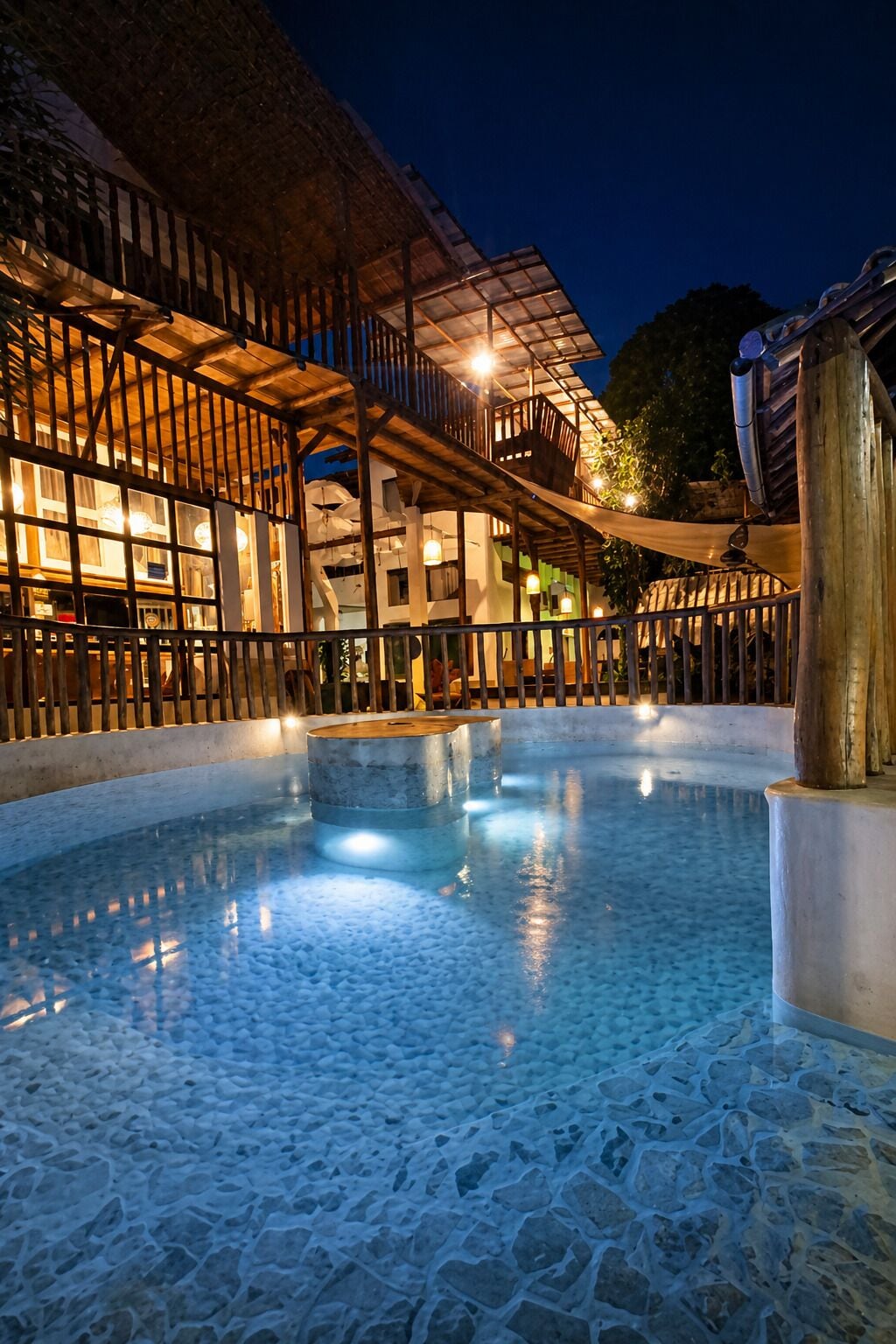 A swimming pool with a stone floor, surrounded by a wooden deck and a building with multiple balconies.