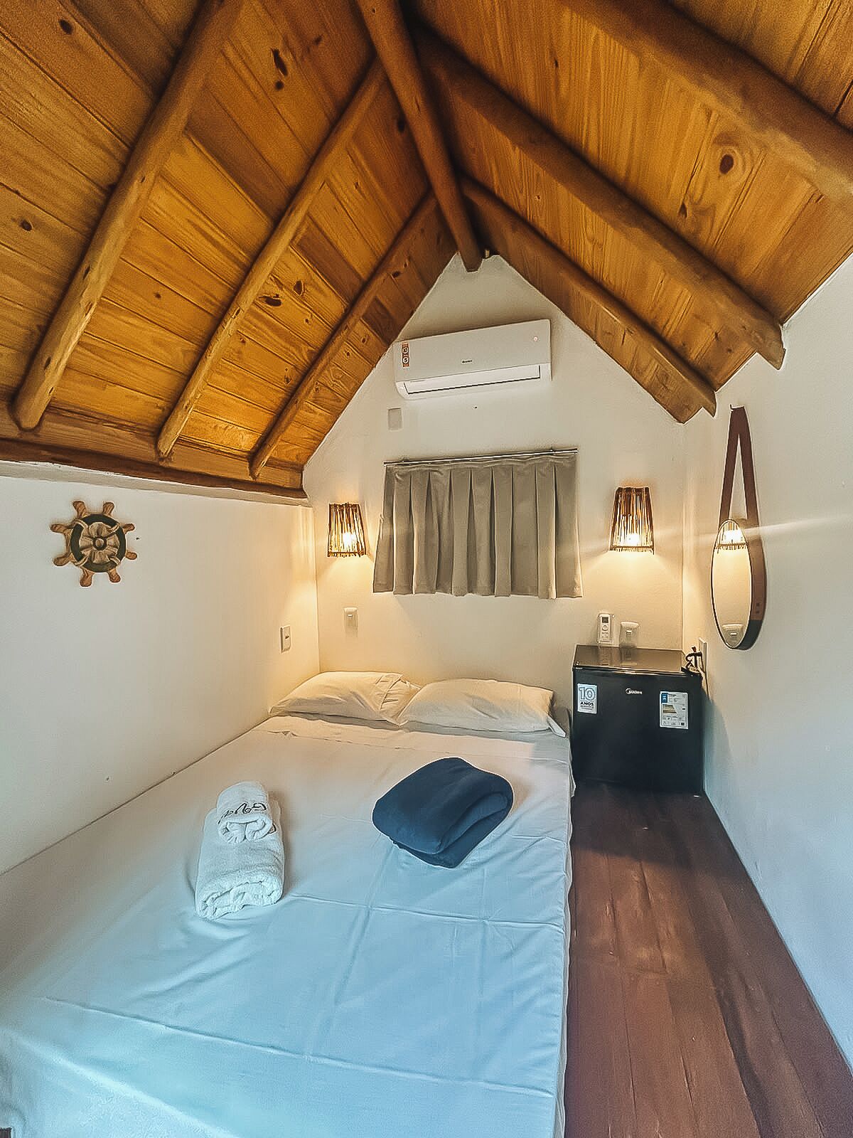 A bedroom with a sloped ceiling, a bed with pillows, a bedside table with a lamp, and a wall-mounted air conditioning unit.