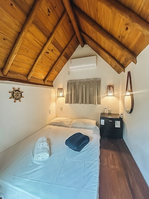 Ein Schlafzimmer mit schrägen Wänden, einem Bett mit Kissen, einem Nachttisch mit Lampe und einer Wand-Klimaanlage.