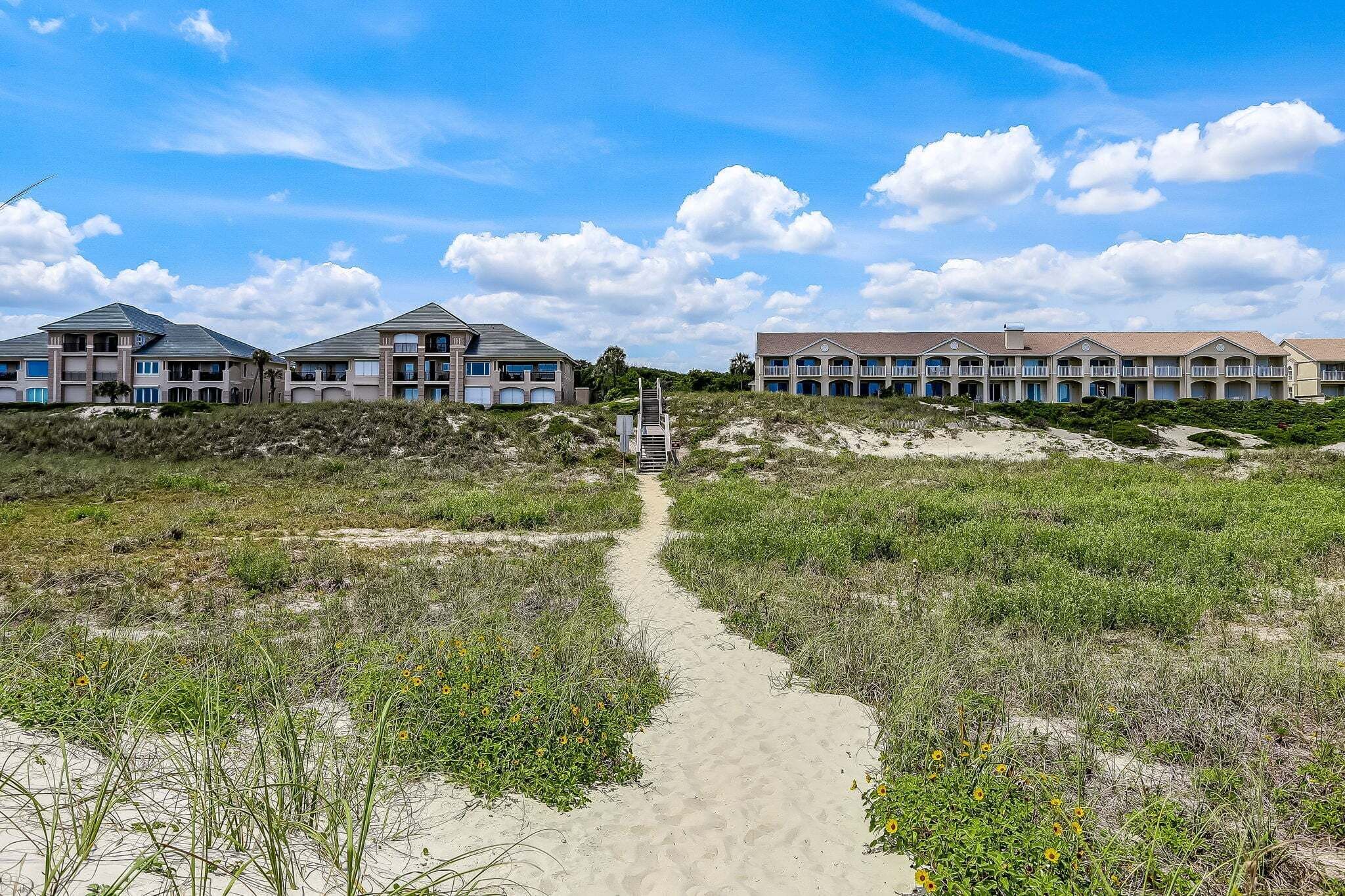 Uma trilha leva a um hotel na praia, com várias varandas e céu claro.