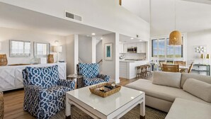 Un salón espacioso con sofá, sillón estampado y una mesa de centro. Se observa una cocina al fondo con armarios blancos y una lámpara colgante.