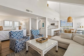 A spacious living area with a sofa, patterned armchair, and a coffee table. A kitchen is visible in the background with white cabinets and a hanging light fixture. - Newly renovated oceanfront multi-level condo inthe Omni Resort Amelia Island (Fernandina Beach)