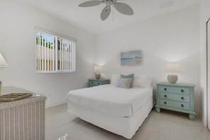 Une chambre à coucher comprenant un lit, deux tables de chevet, un ventilateur de plafond, une fenêtre avec des stores et un tableau accroché au mur.