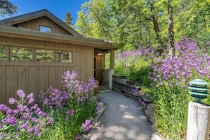 A wooden cabin with a gravel path, surrounded by purple flowers and trees.