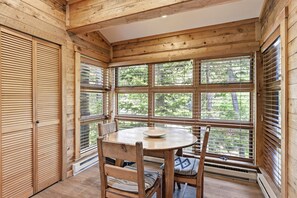 Un intérieur en bois avec une table ronde et des chaises, de grandes fenêtres équipées de stores et une vue sur la verdure à l’extérieur.