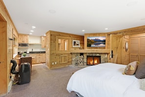 A room with a fireplace, wooden walls, a bed, a desk with a computer, and a kitchen area.