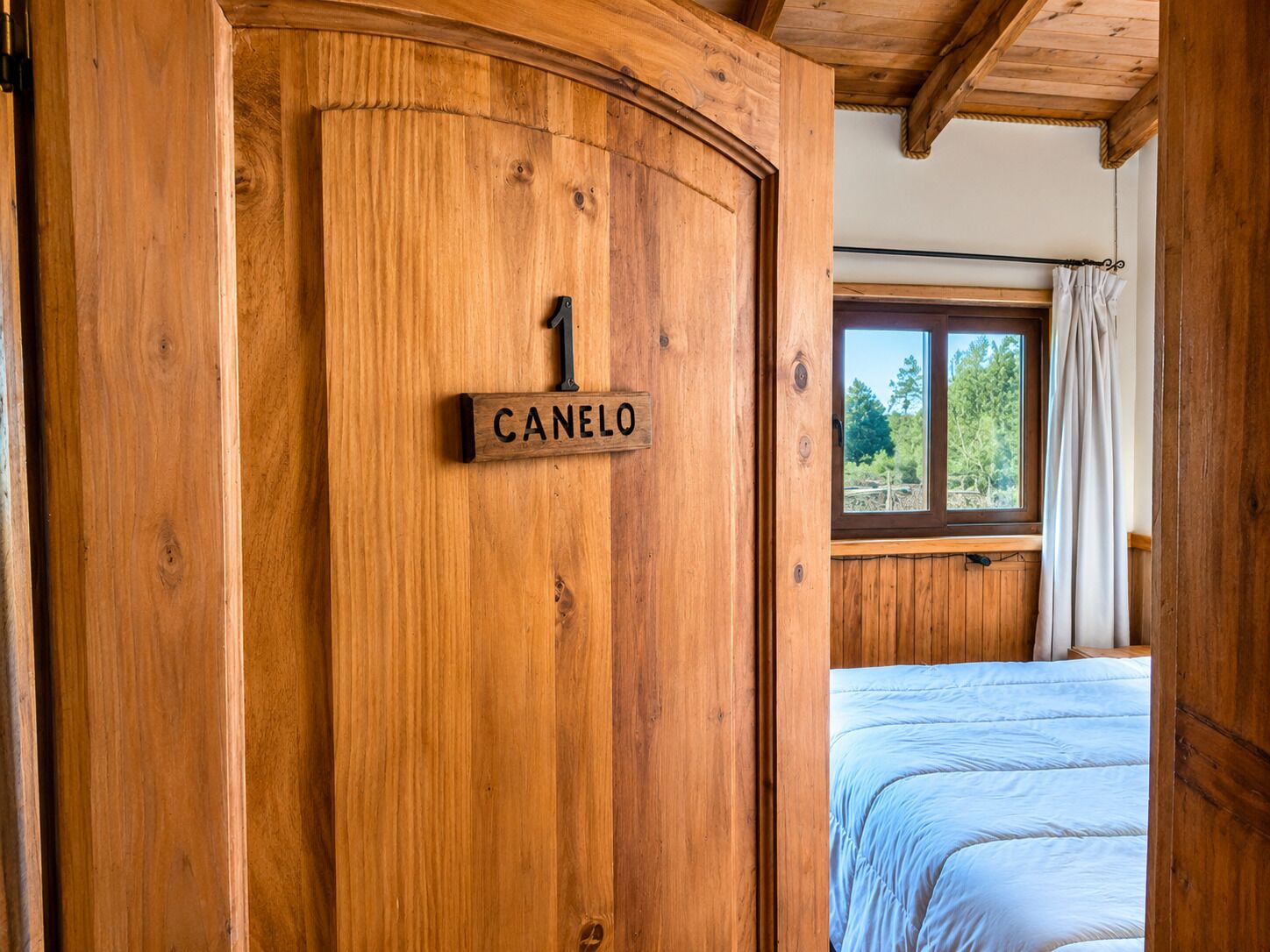 Eine Holztür mit einem Schild, auf dem '1 CANELO' steht, ein Fenster mit Vorhängen und ein Bett mit weißer Daunendecke.