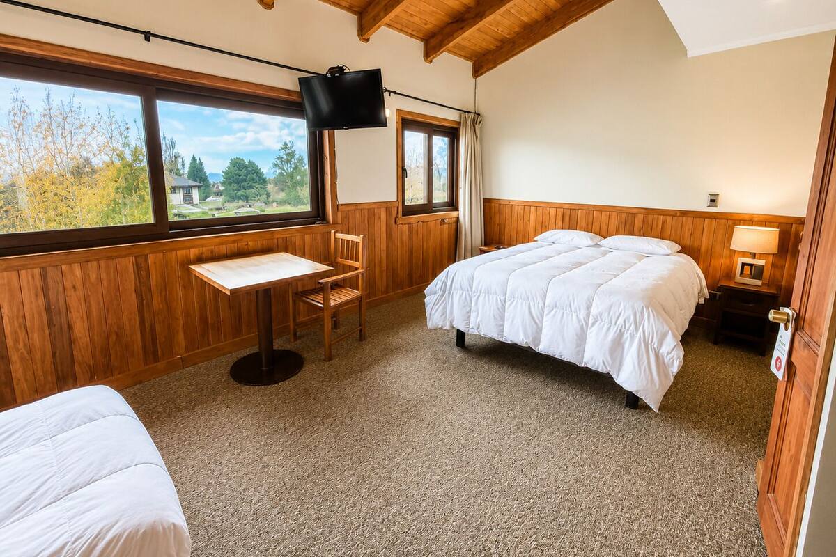 Un dormitorio con cama, escritorio, silla, televisor y ventana con vista a árboles.