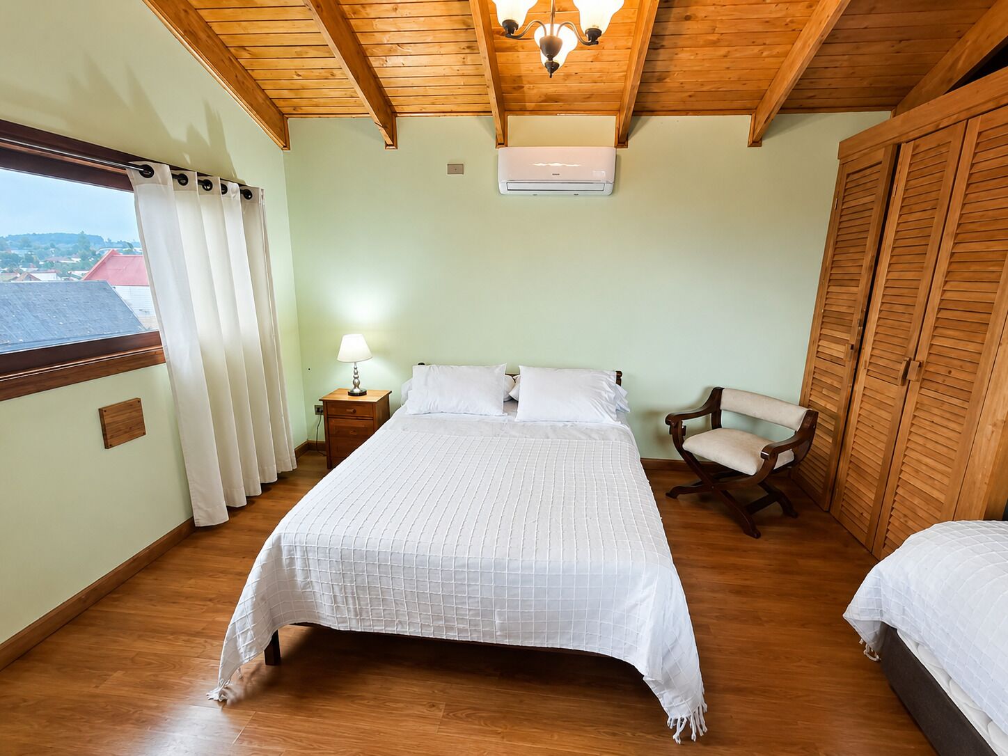 A bedroom with a bed, a chair, a nightstand, an air conditioner, and a window with curtains.