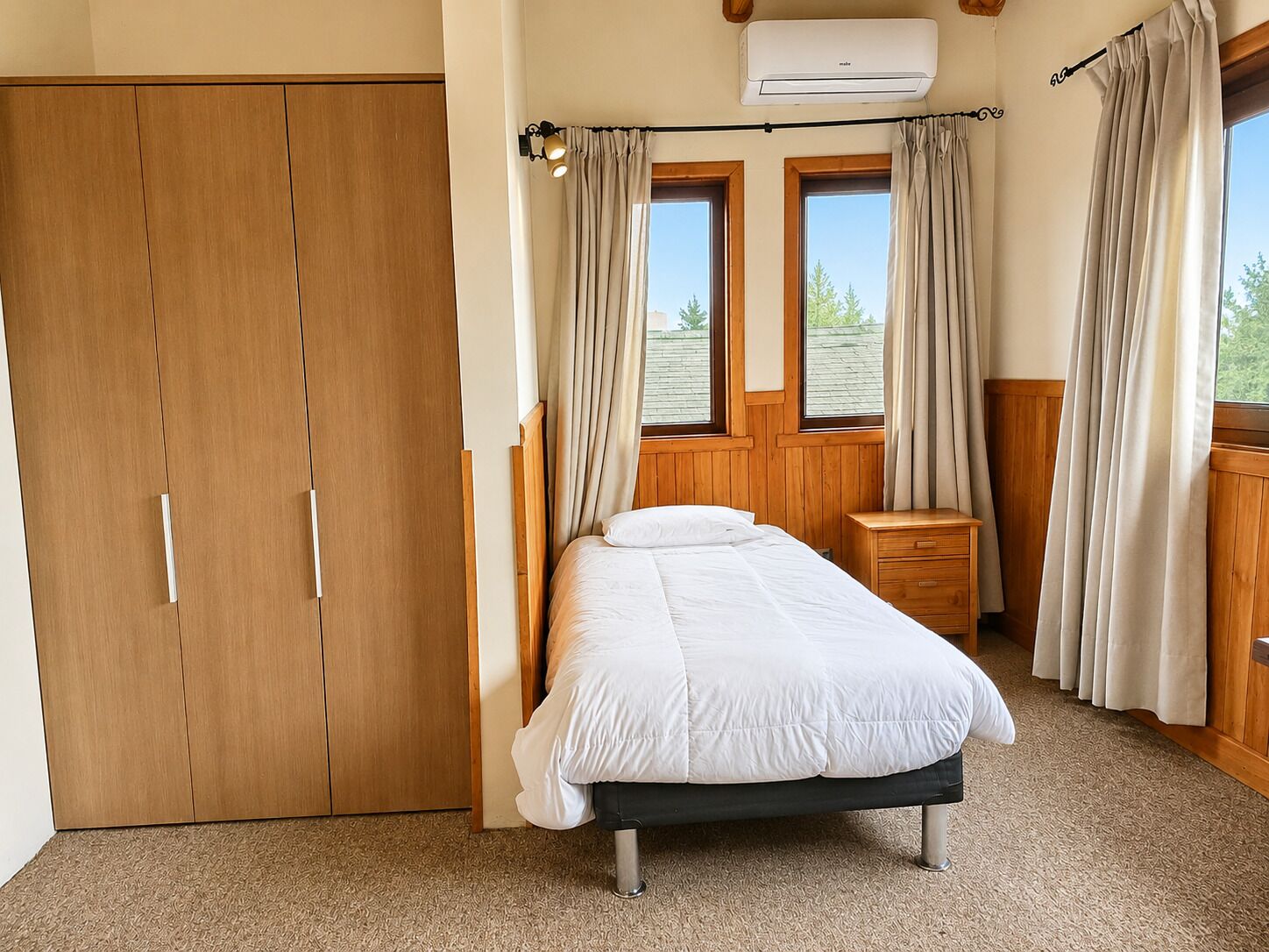 Een slaapkamer met een bed, een houten garderobe, twee ramen met gordijnen, een nachtkastje en een airconditioning.