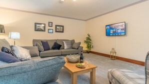 Una sala de estar con un sofá gris, una mesa de centro de madera con una botella y vasos, un televisor en la pared y cuadros enmarcados.