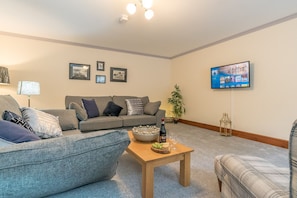 Ein Wohnzimmer mit grauem Sofa, einem Holztisch mit Flasche und Gläsern, einem Fernseher an der Wand und gerahmten Bildern.