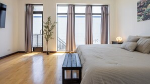 Quarto com uma cama grande, um banco de madeira, uma planta em vaso, uma mesinha de cabeceira com abajur e vista para uma varanda.