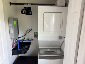 A compact laundry room with a stacked washer and dryer, a thermostat, and a small shelf with cleaning supplies.